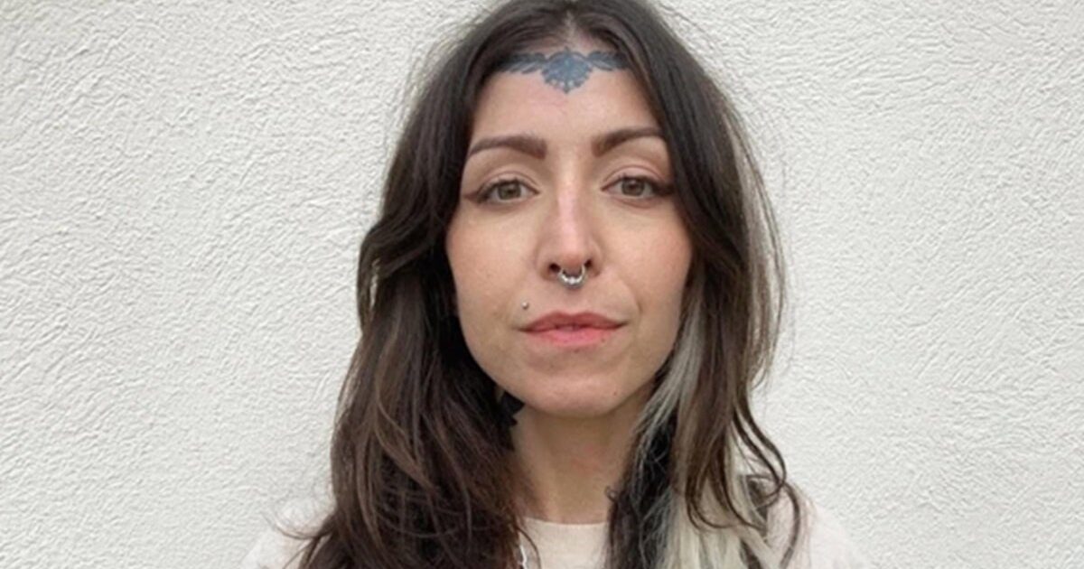 Portrait of a woman with long dark hair, facial piercings, and a decorative tattoo on her forehead, set against a textured white wall.