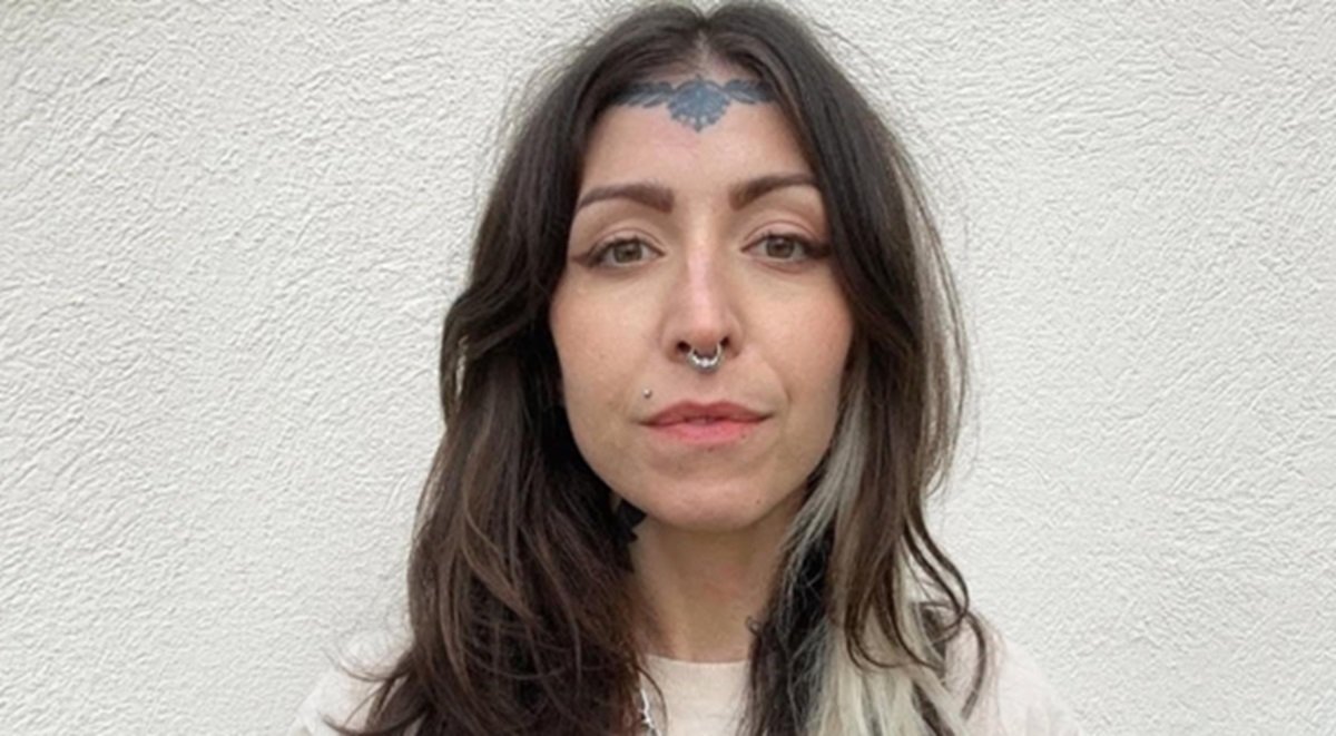 Portrait of a woman with long dark hair, facial piercings, and a decorative tattoo on her forehead, set against a textured white wall.