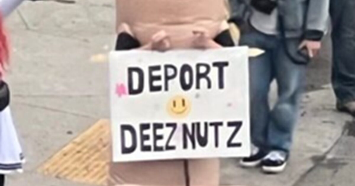 Costumed performer holding a humorous sign reading "Deport Deez Nutz" at a public event, surrounded by onlookers.