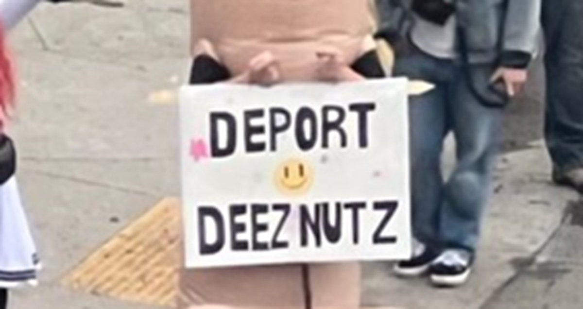 Costumed performer holding a humorous sign reading "Deport Deez Nutz" at a public event, surrounded by onlookers.