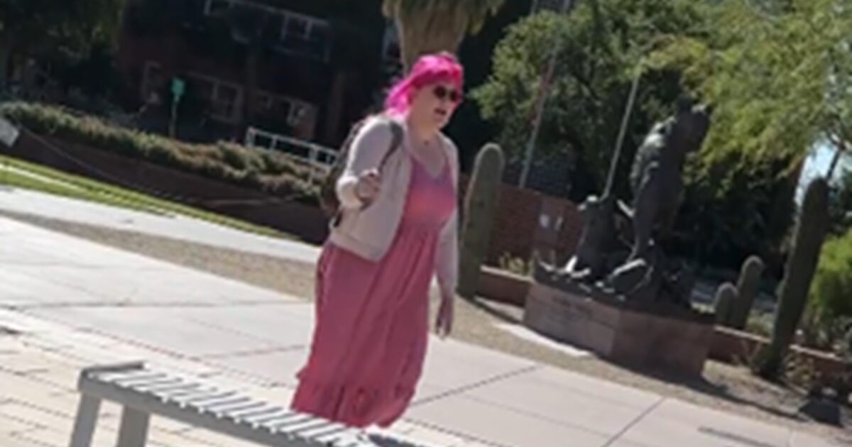 Woman with pink hair wearing a pink dress and sunglasses stands outdoors near a statue in a park setting.