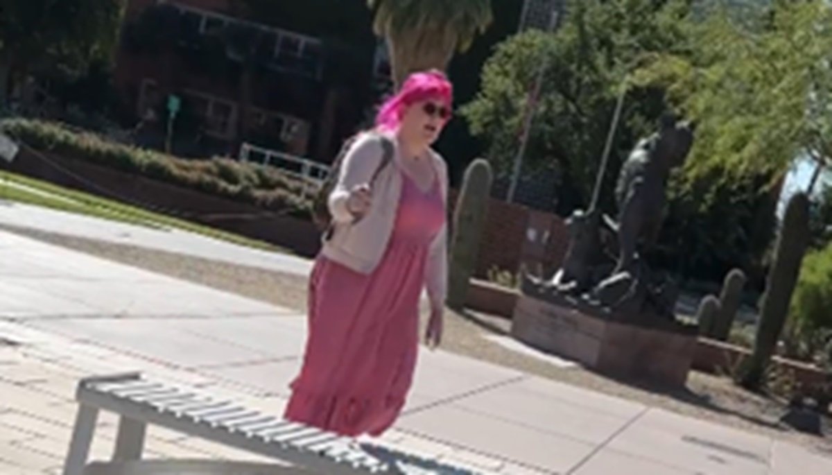 Woman with pink hair wearing a pink dress and sunglasses stands outdoors near a statue in a park setting.