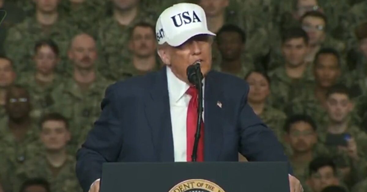 Donald Trump speaking at a military event, wearing a USA hat, with a crowd of soldiers in the background.