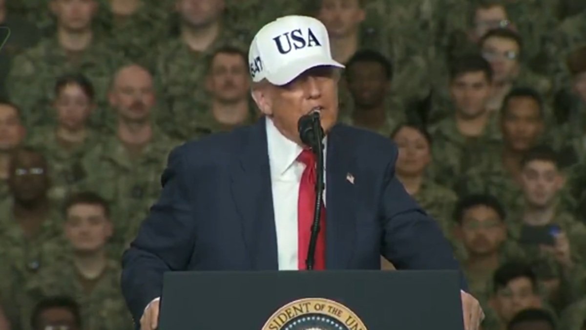 Donald Trump speaking at a military event, wearing a USA hat, with a crowd of soldiers in the background.