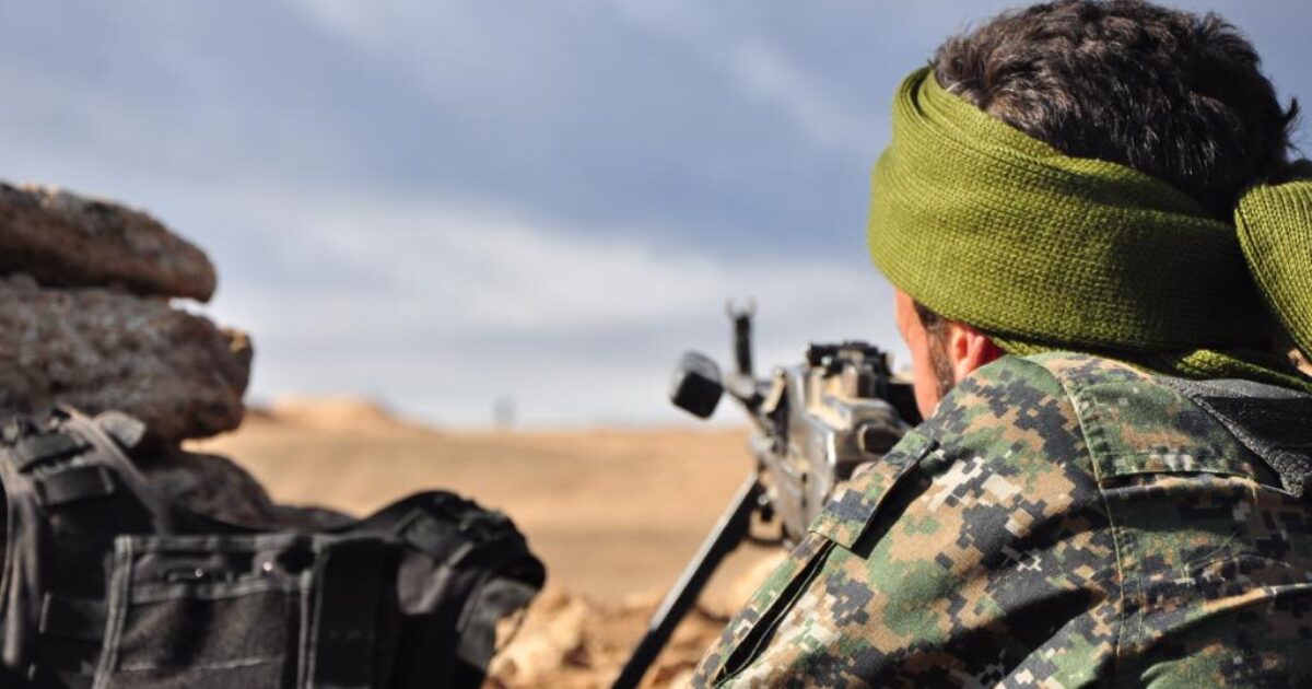 Soldier in camouflage gear and green headscarf aiming a rifle from a fortified position in a desert landscape.
