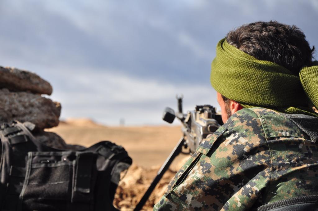 Soldier in camouflage gear and green headscarf aiming a rifle from a fortified position in a desert landscape.