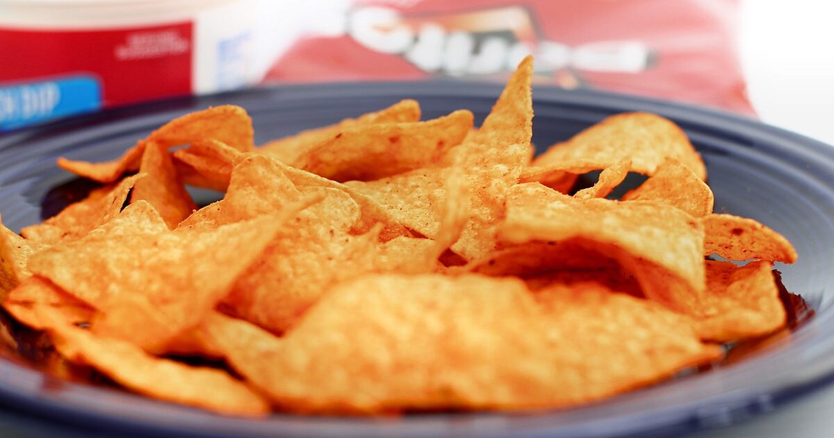 Close-up of crispy tortilla chips on a blue plate, with a container of dip and a bag of chips in the background.
