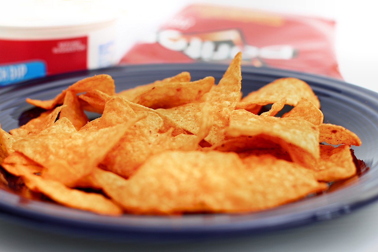 Close-up of crispy tortilla chips on a blue plate, with a container of dip and a bag of chips in the background.