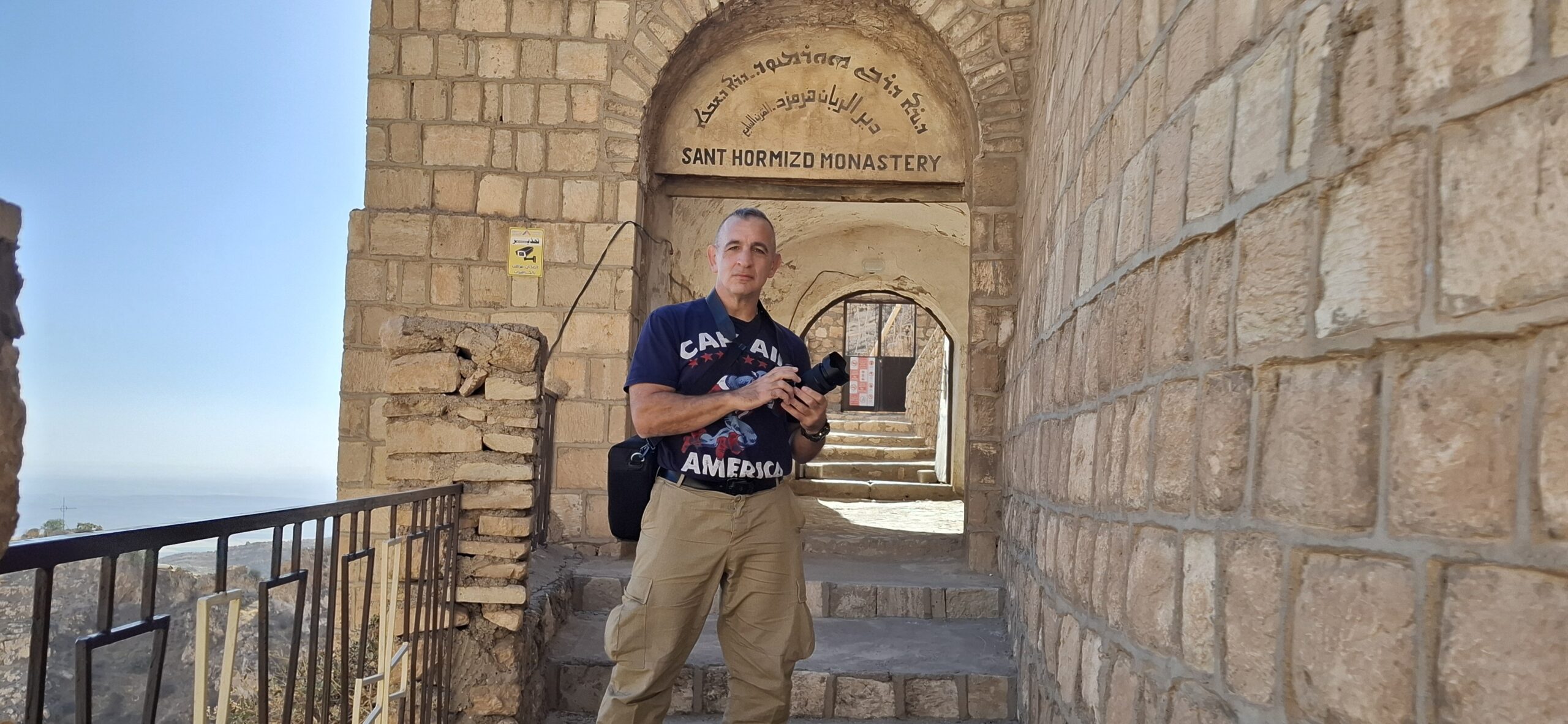 Man holding a camera stands at the entrance of Saint Hormizd Monastery, showcasing ancient stone architecture and a clear sky in the background.