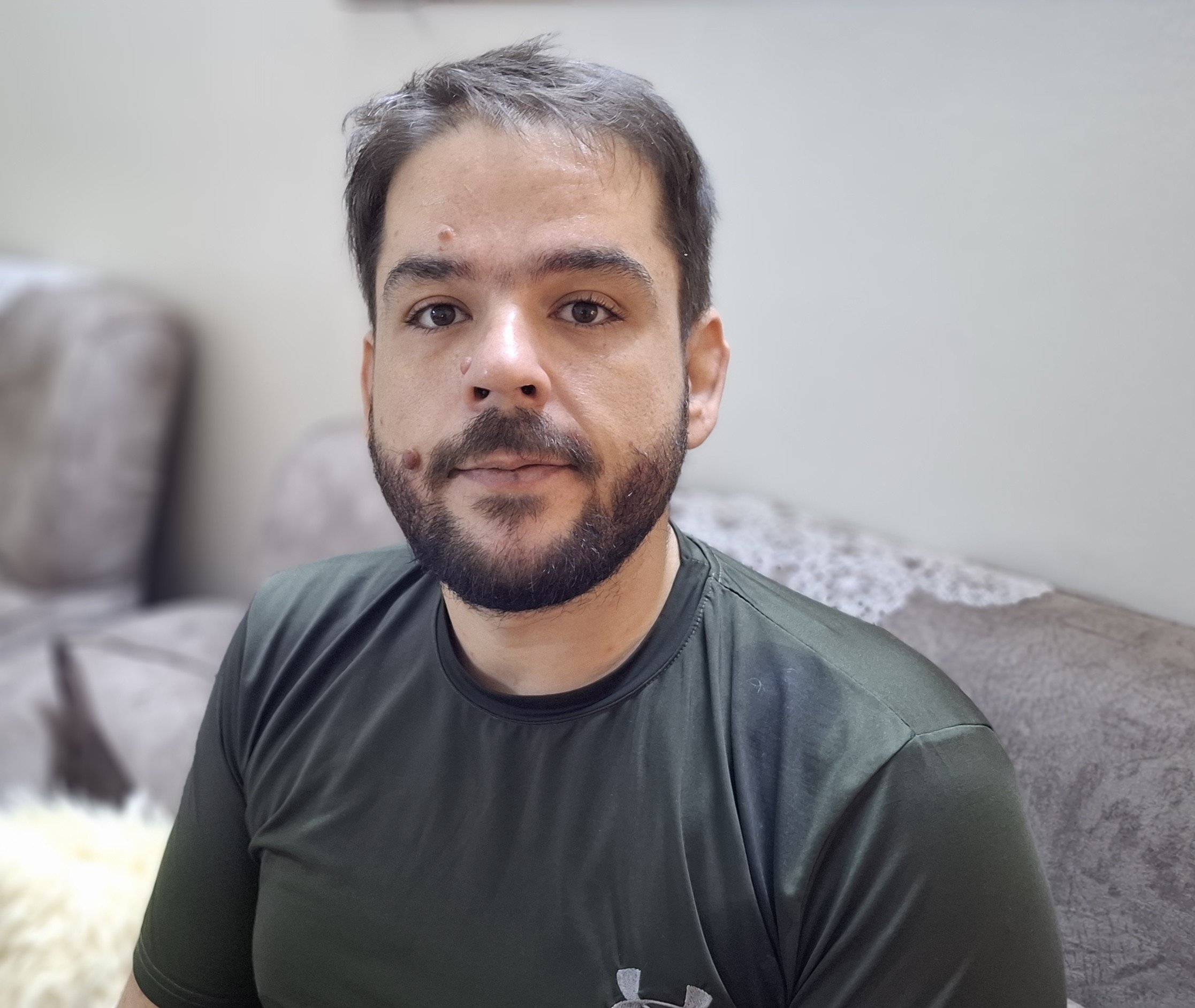 Man with a beard wearing a green athletic shirt sits on a couch, looking directly at the camera in a well-lit indoor setting.