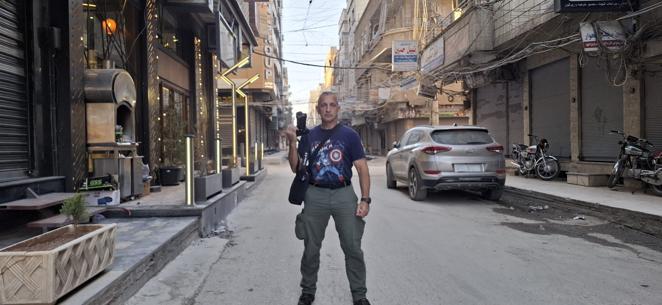 A person stands confidently on a quiet street, showcasing a vibrant t-shirt and holding a camera, surrounded by shops and parked vehicles.
