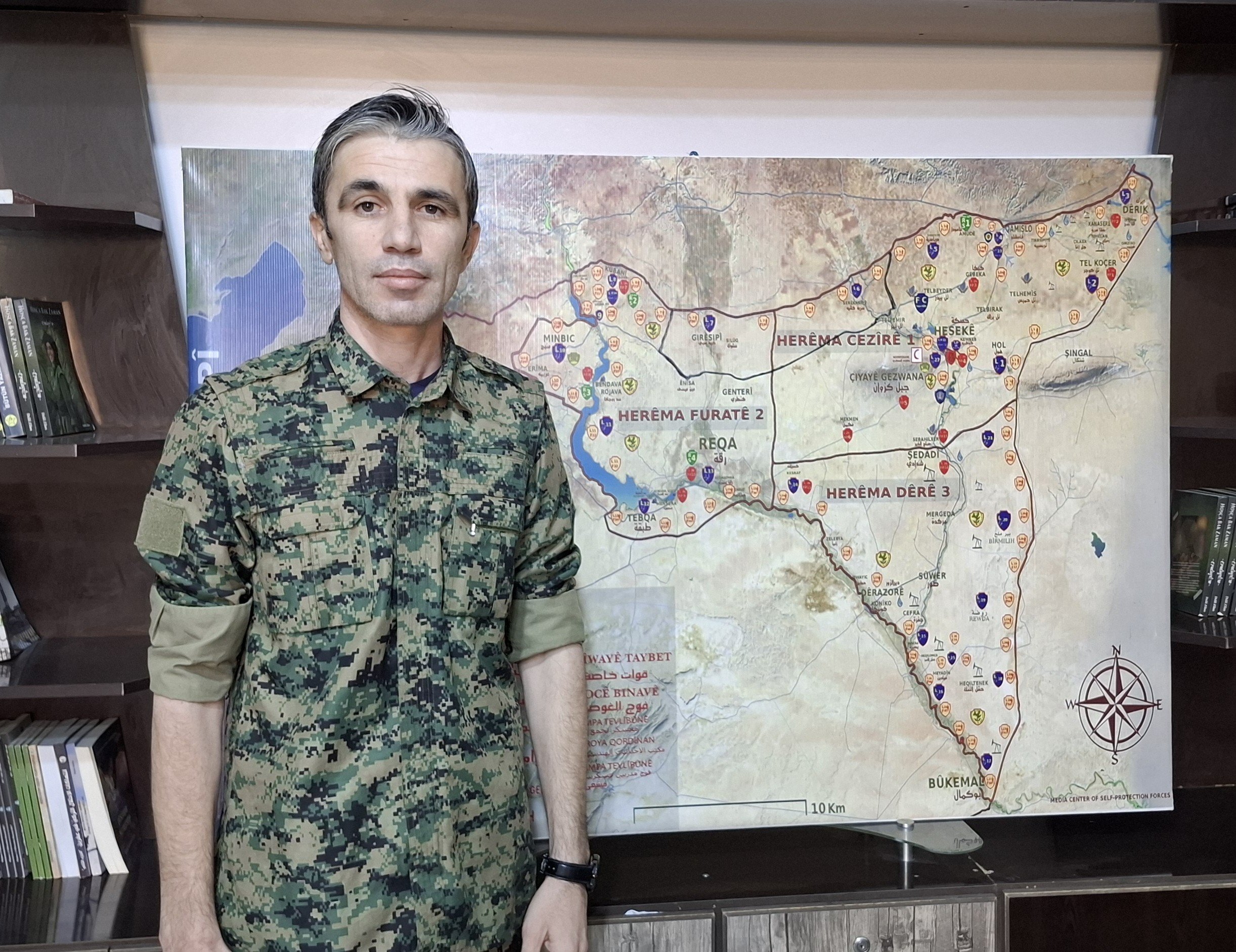 Man in military camouflage stands in front of a detailed map of regions, highlighting strategic locations and symbols, with bookshelves in the background.