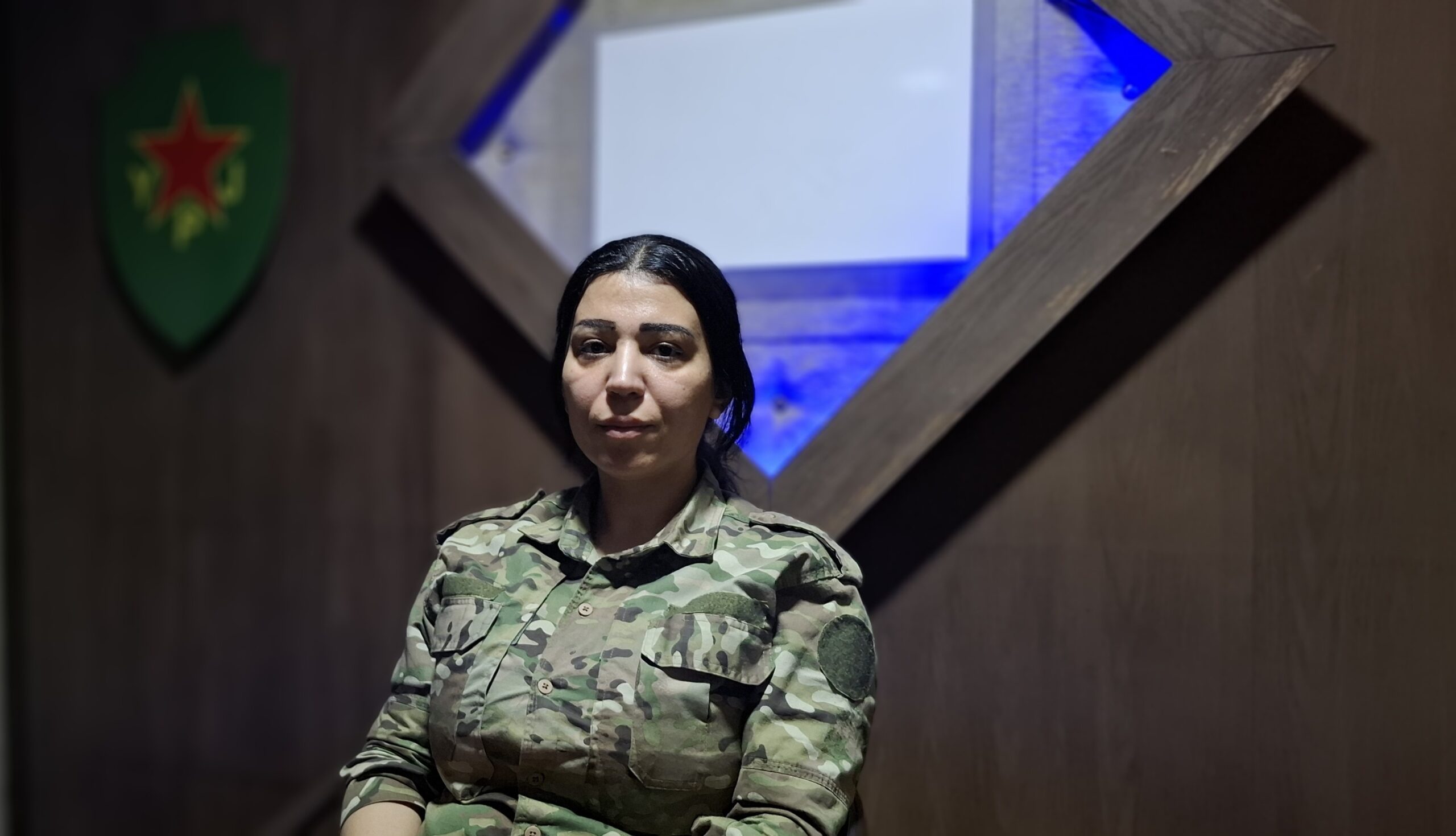 Female soldier in camouflage uniform seated against a wooden backdrop with blue lighting, representing military strength and dedication.