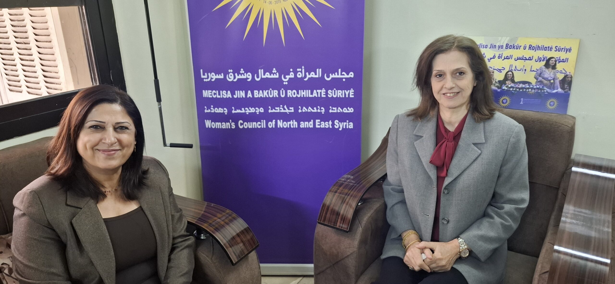 Two women seated in front of a banner for the Woman's Council of North and East Syria, discussing women's rights and empowerment initiatives.