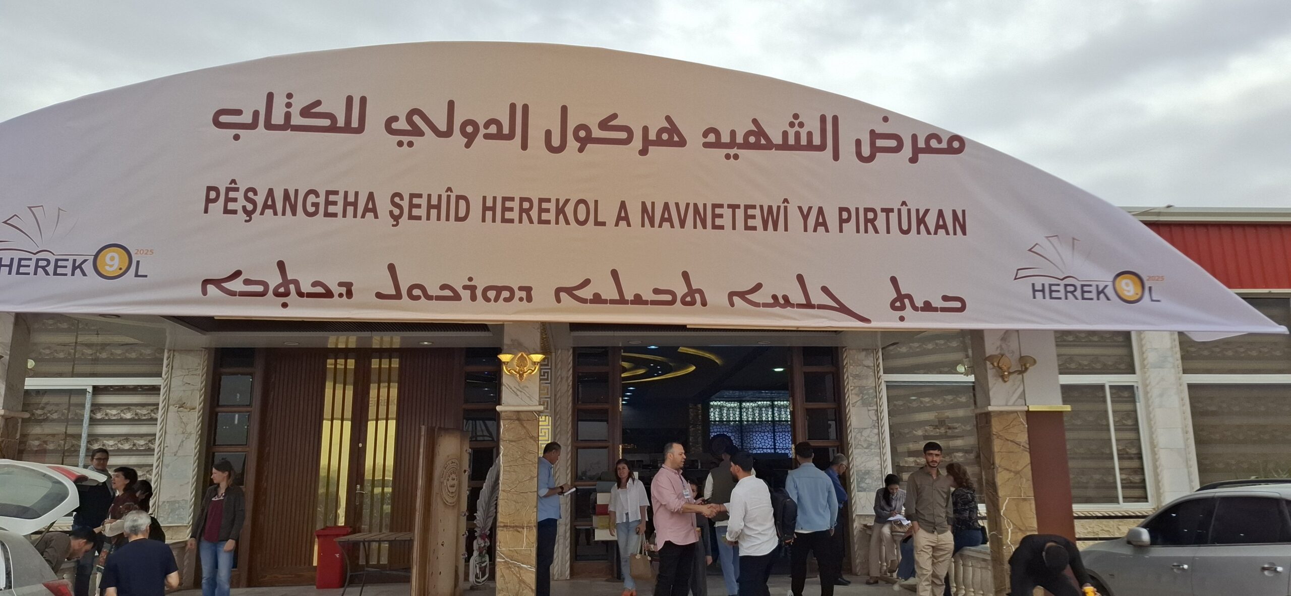Entrance of the Herekol International Book Fair, featuring a welcoming banner in multiple languages, with attendees interacting outside the venue.