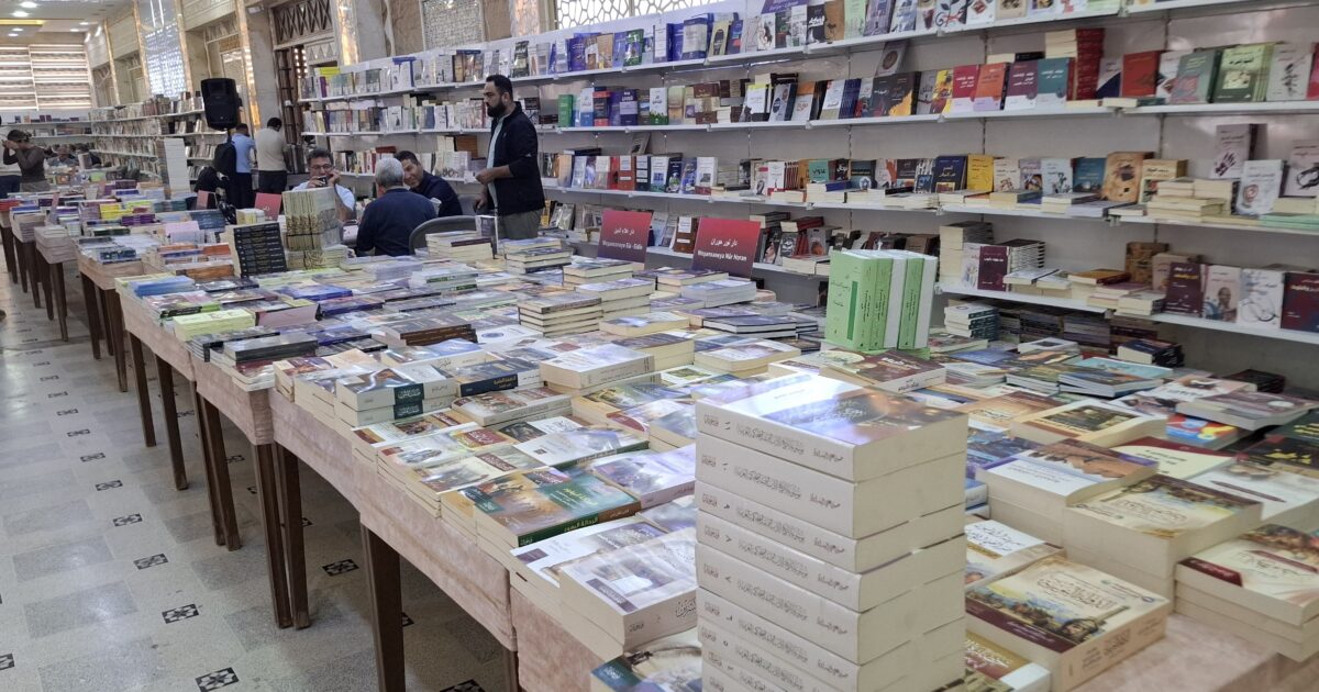 A busy book fair with tables filled with various books, people browsing, and shelves lined with literature in a well-lit space.