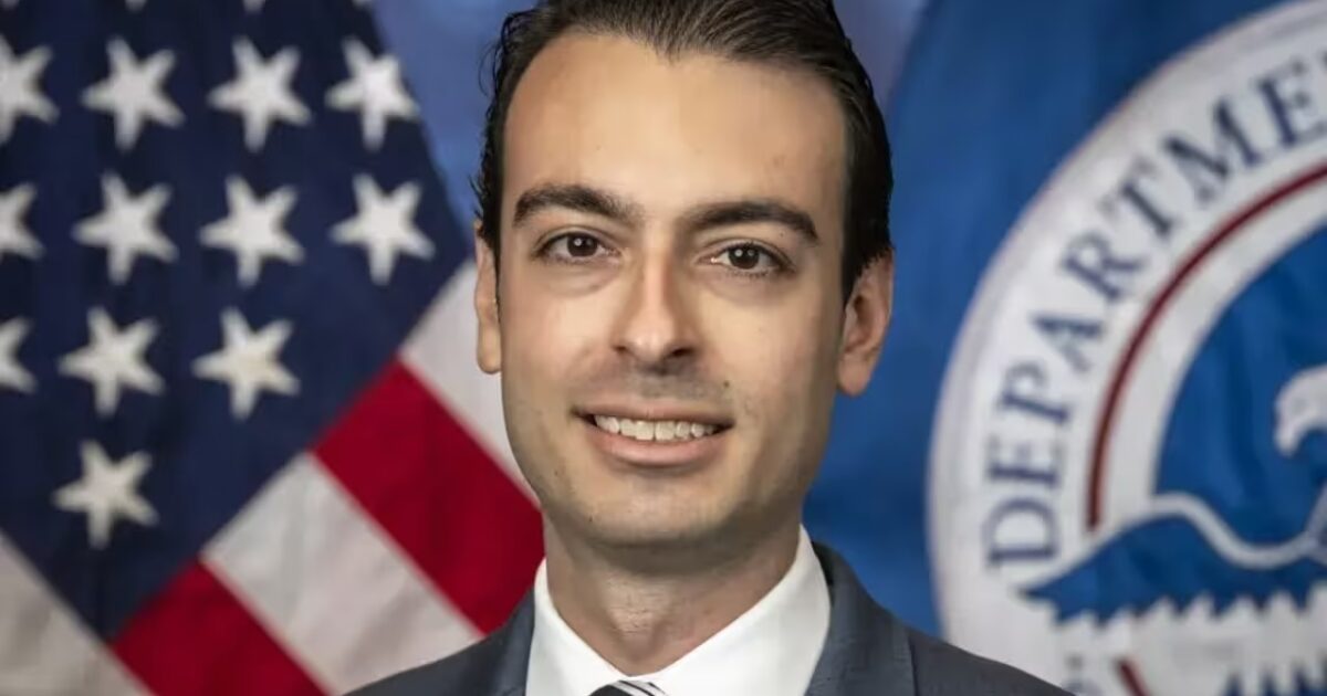 Professional headshot of a government official with a blue suit and tie, smiling against a backdrop of the American flag and Department of Homeland Security seal.