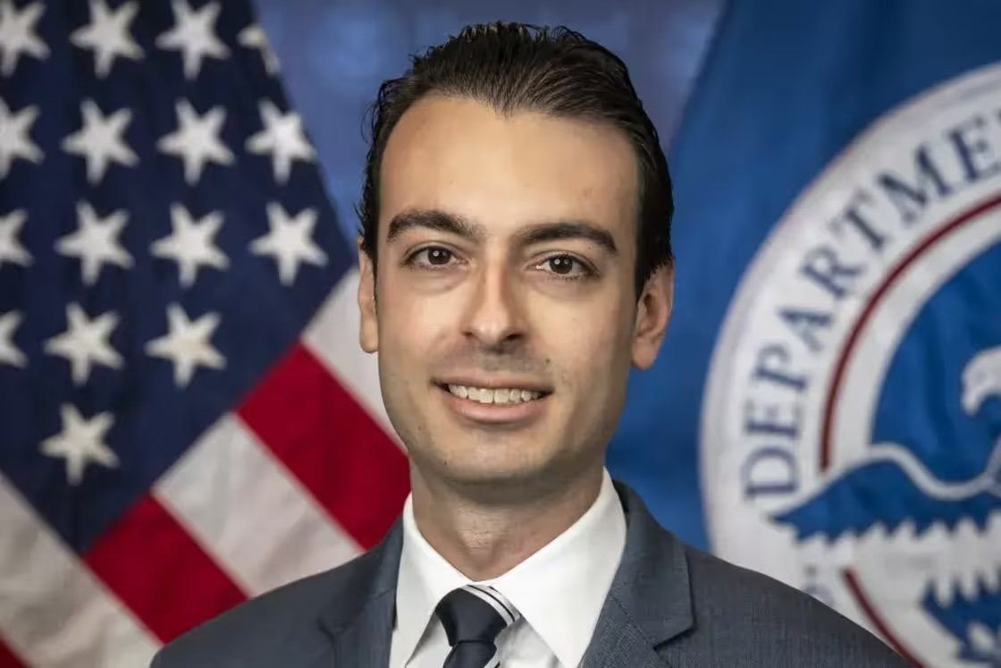 Professional headshot of a government official with a blue suit and tie, smiling against a backdrop of the American flag and Department of Homeland Security seal.