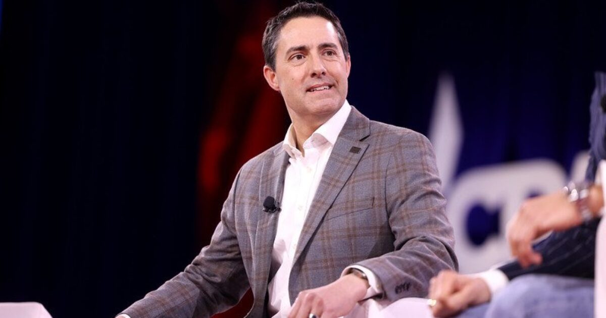 Business professional speaking at a conference panel, wearing a gray blazer, with a focused expression and a microphone attached, set against a colorful backdrop.