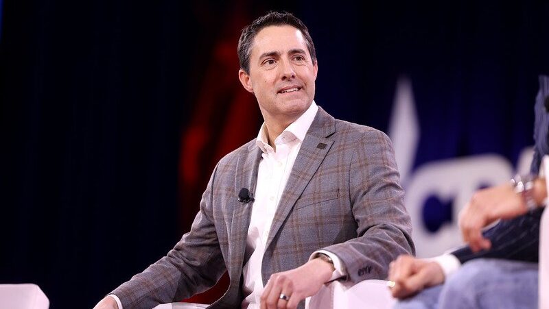 Business professional speaking at a conference panel, wearing a gray blazer, with a focused expression and a microphone attached, set against a colorful backdrop.