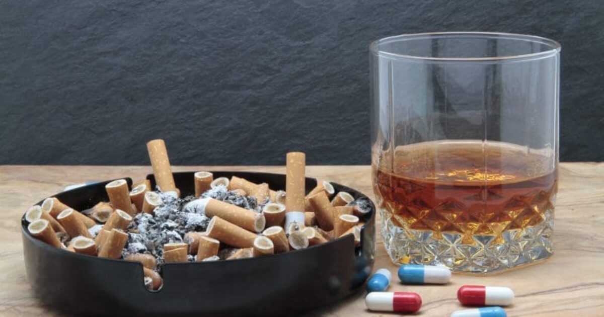 Ashtray filled with cigarette butts, a glass of whiskey, and scattered capsules on a wooden surface, symbolizing smoking and substance use.