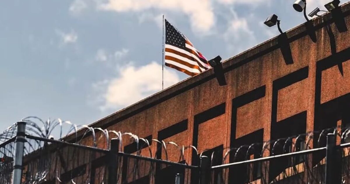 American flag flying above a prison building surrounded by barbed wire and security cameras under a blue sky.