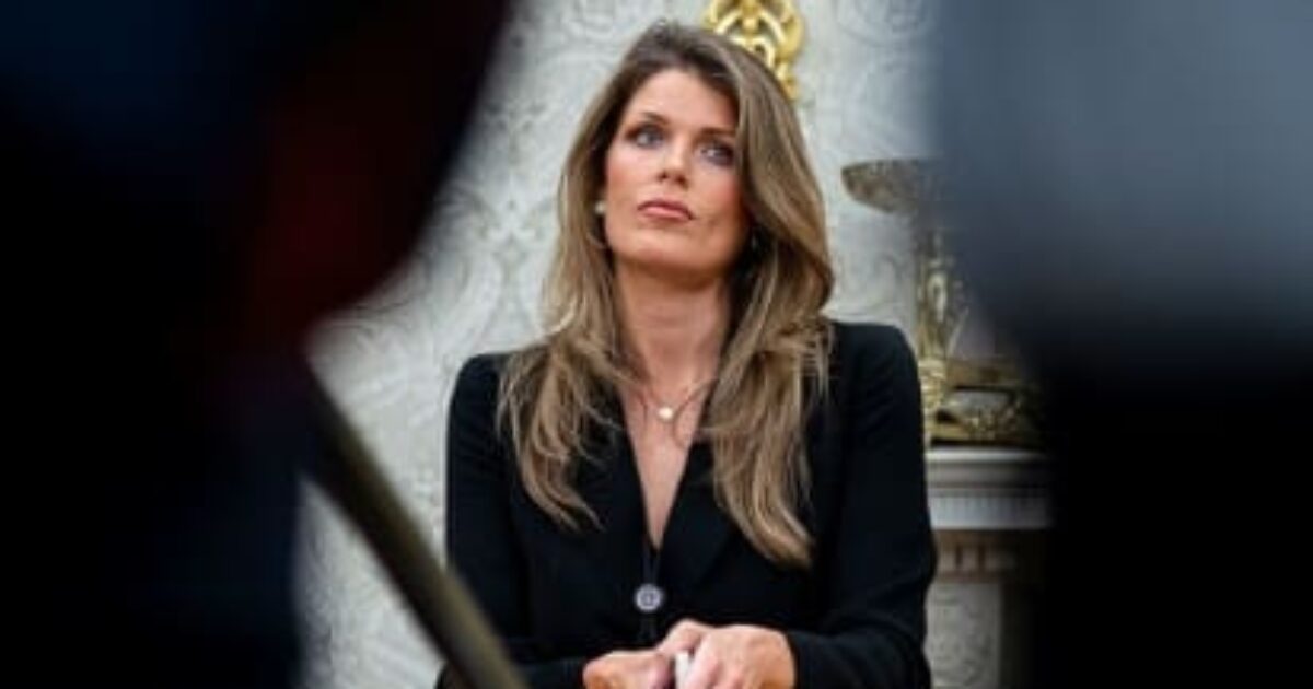 A woman with long hair and a black blazer sits thoughtfully in a formal setting, surrounded by blurred figures.