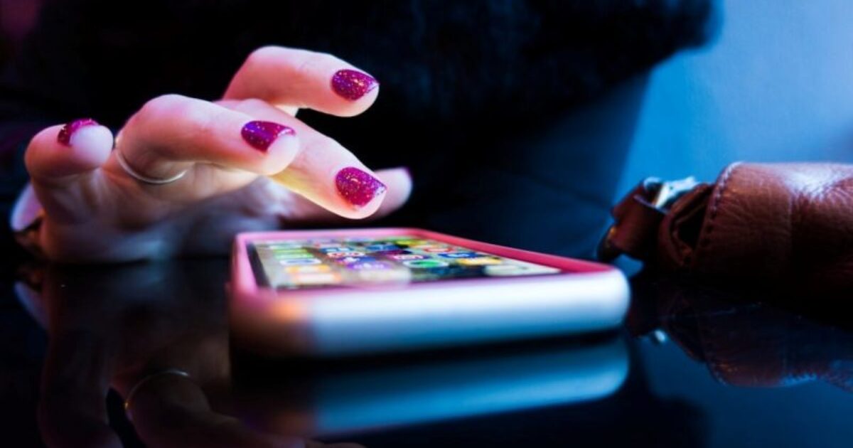 Close-up of a hand with glittery nails tapping on a smartphone screen, reflecting on a glossy black surface.