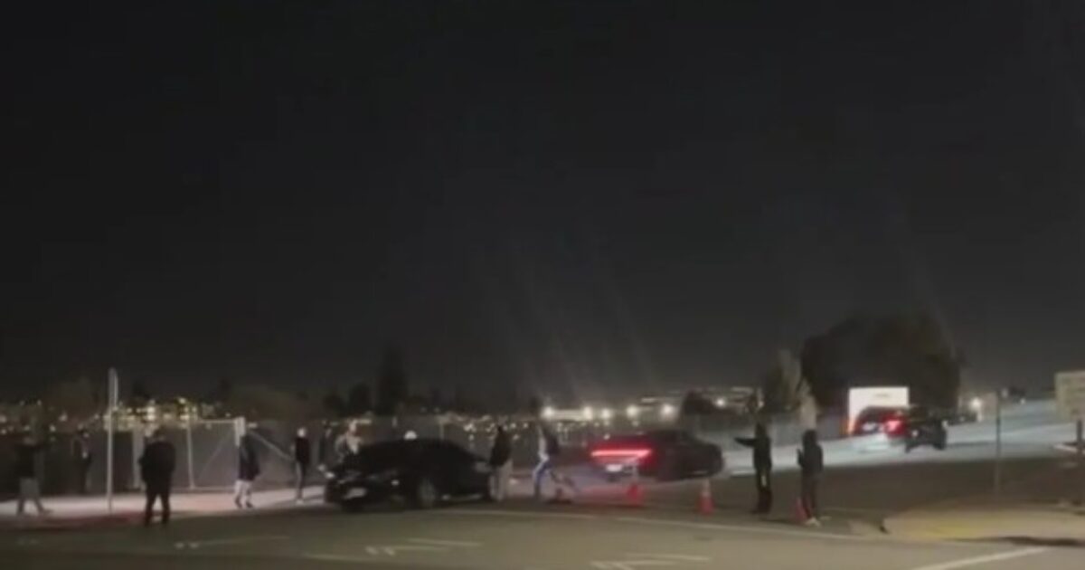 Crowd gathered at night near parked cars on a dimly lit street, with city lights visible in the background.