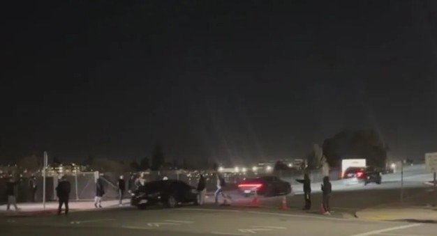 Crowd gathered at night near parked cars on a dimly lit street, with city lights visible in the background.