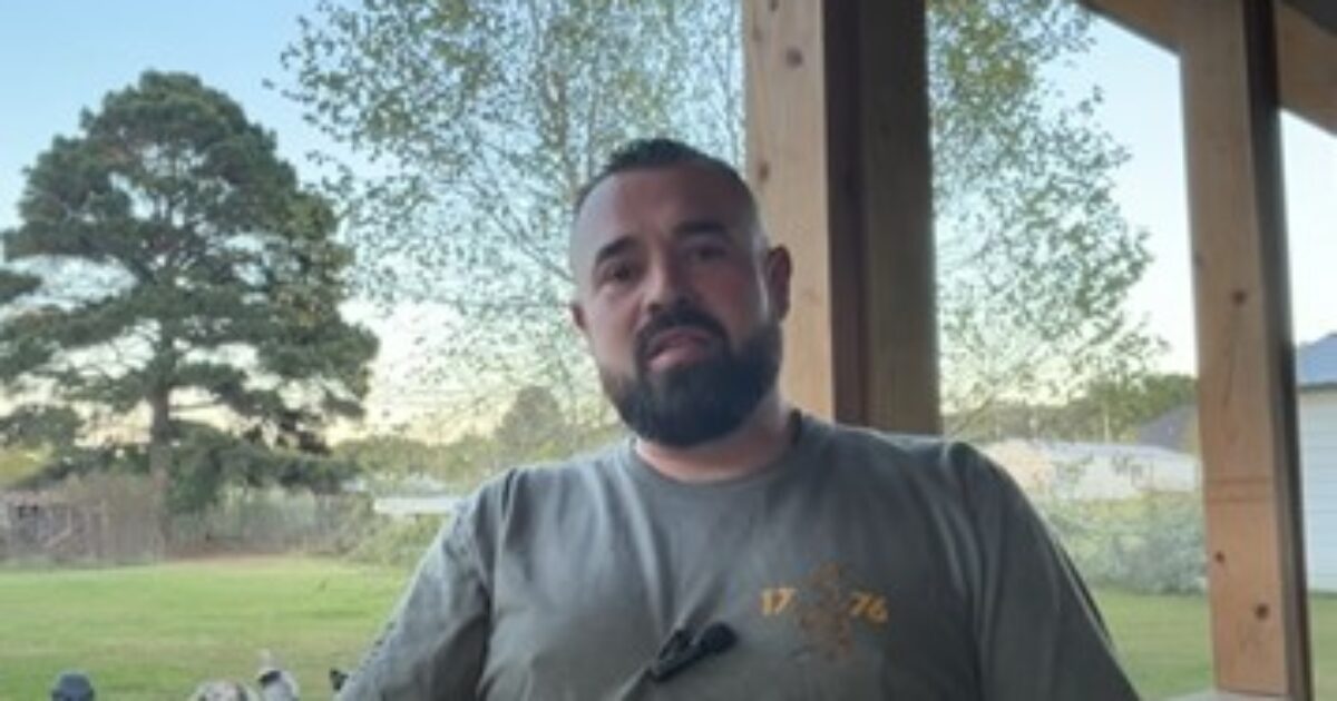 Man with a beard sitting outdoors on a porch, surrounded by greenery and a view of a yard with trees.