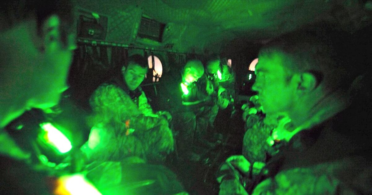 Soldiers seated inside a military aircraft, illuminated by green lights, preparing for a night operation.