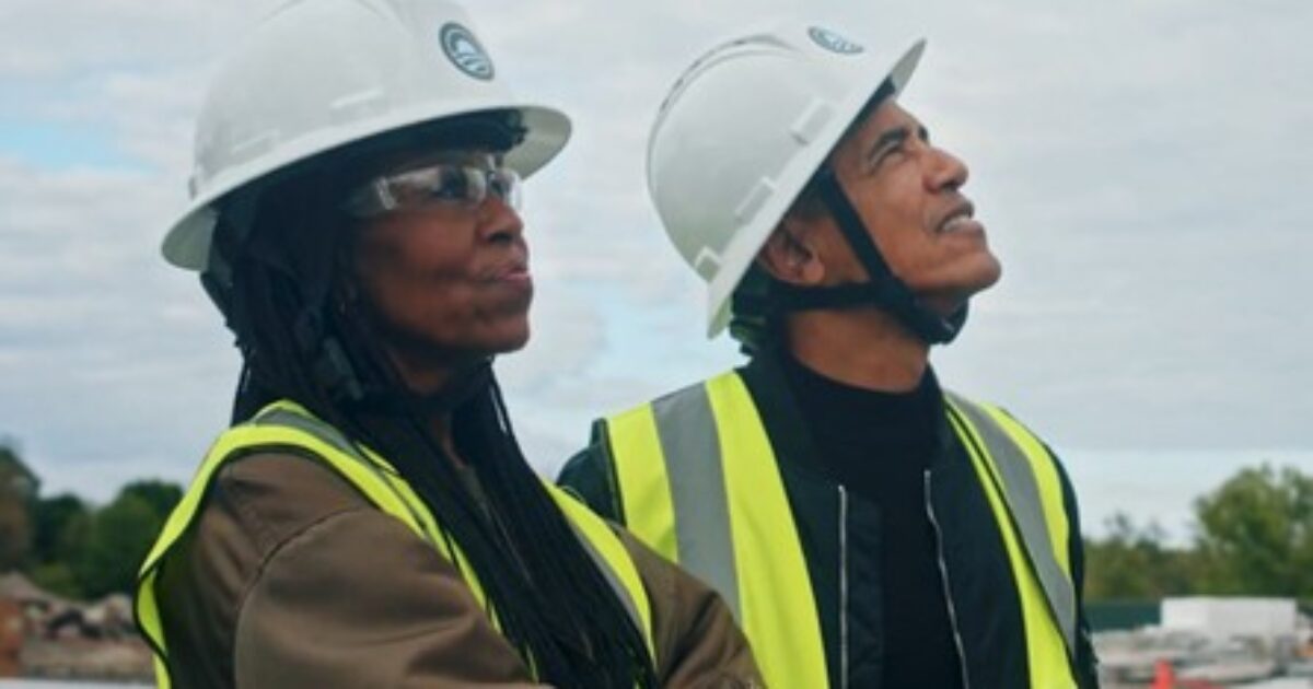 Two construction workers in hard hats and safety vests looking upwards, observing a building site with a cloudy sky in the background.