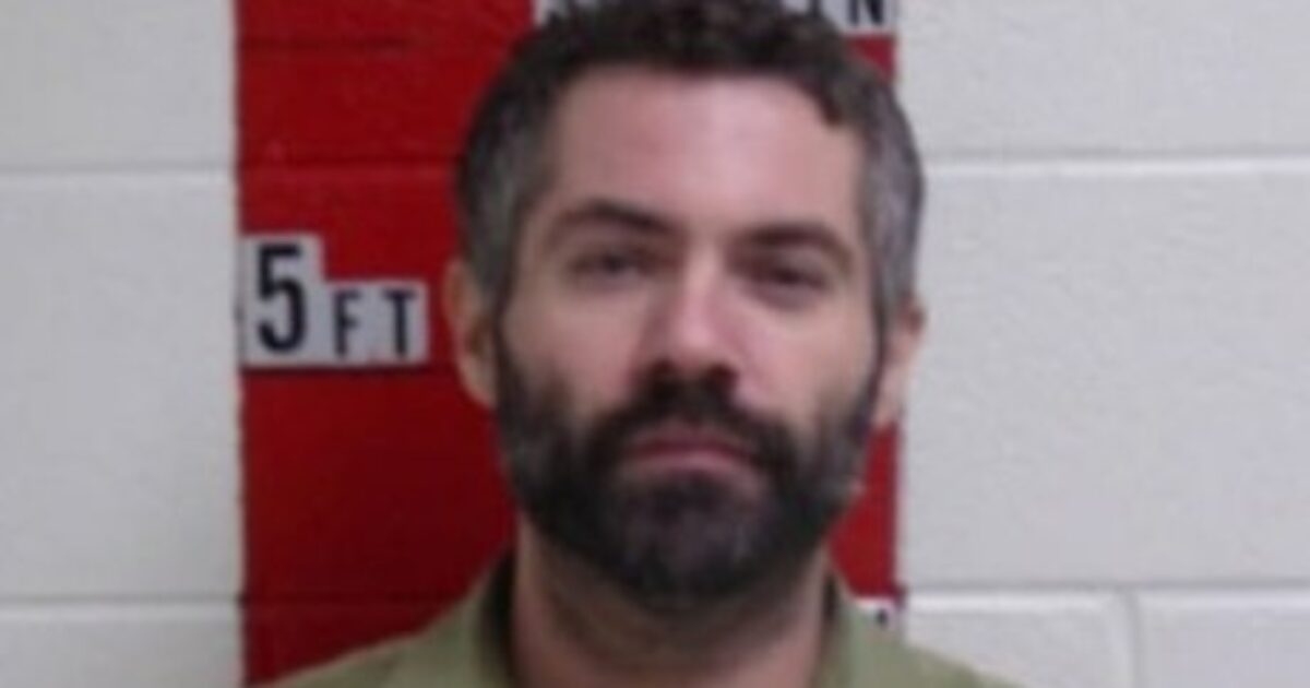 Mugshot of a man with a beard, wearing a green shirt, standing against a height measurement chart on a wall.