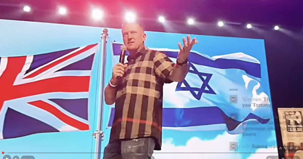 Tommy Robinson speaking on stage with British and Israeli flags in the background, addressing an audience at a public event.