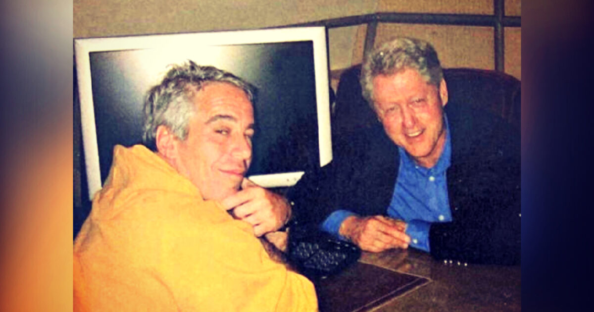 Two men posing together at a desk with a computer in the background, one wearing an orange hoodie and the other in a black jacket and blue shirt.