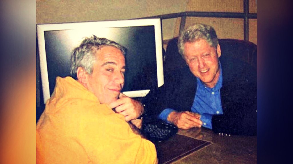 Two men posing together at a desk with a computer in the background, one wearing an orange hoodie and the other in a black jacket and blue shirt.