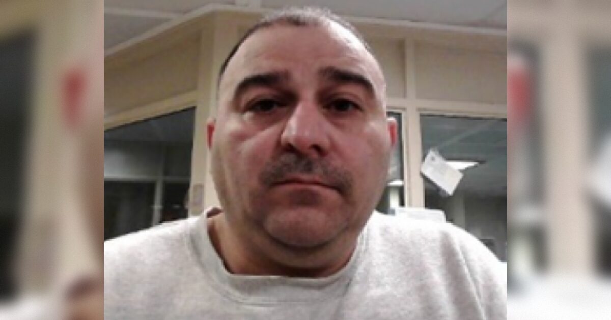 Close-up portrait of a man with short hair and a mustache, wearing a gray sweatshirt, seated in an indoor setting with blurred background elements.
