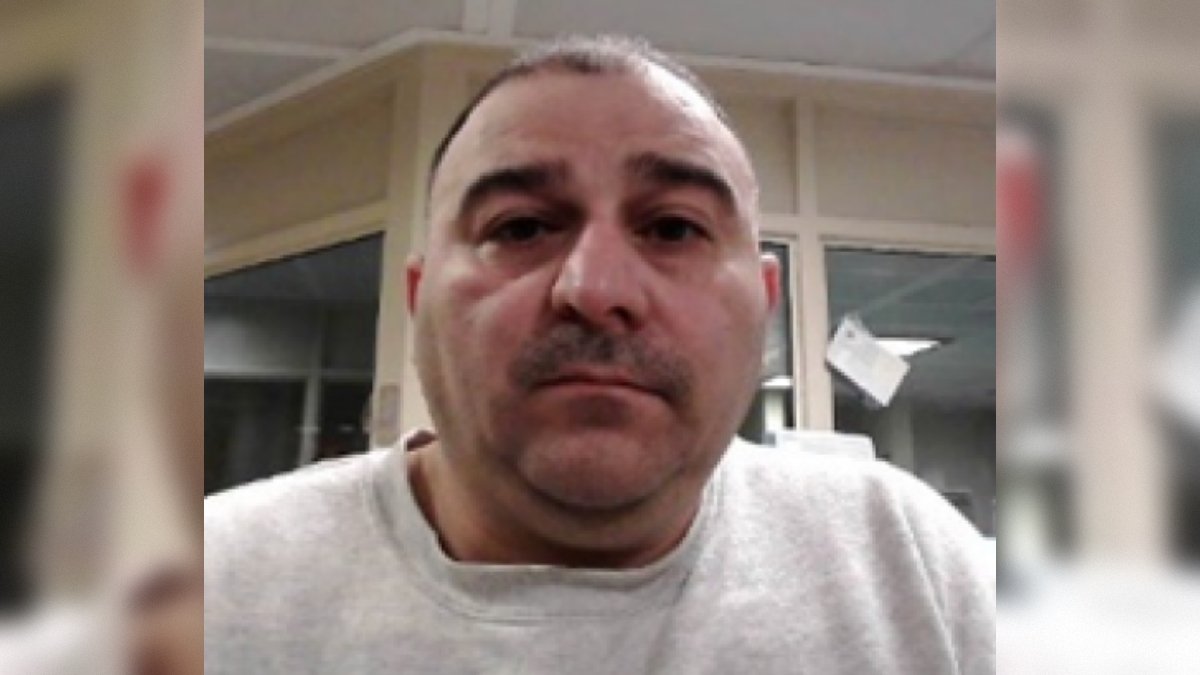 Close-up portrait of a man with short hair and a mustache, wearing a gray sweatshirt, seated in an indoor setting with blurred background elements.