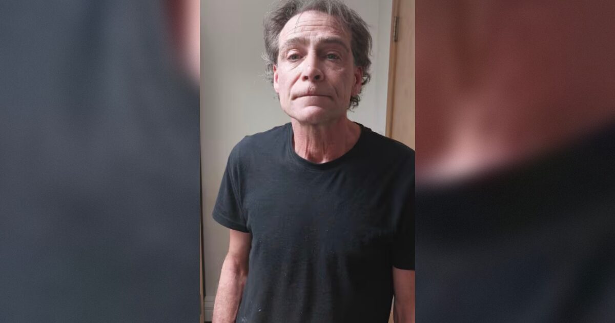 Man with disheveled hair wearing a black t-shirt stands against a neutral background, looking directly at the camera.