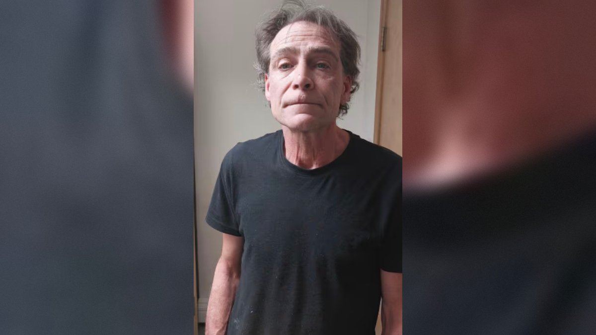 Man with disheveled hair wearing a black t-shirt stands against a neutral background, looking directly at the camera.