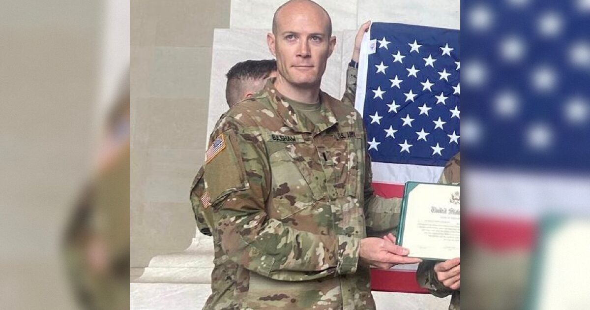 Service member in military uniform holding a certificate and standing in front of an American flag during a formal event.