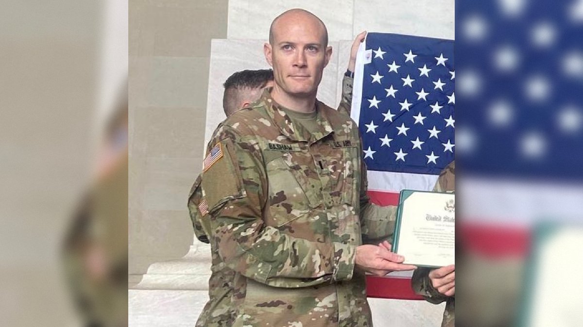Service member in military uniform holding a certificate and standing in front of an American flag during a formal event.