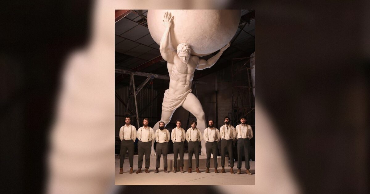 A group of men in vintage attire stands in front of a large statue of Atlas holding up the world, showcasing a blend of art and fashion.