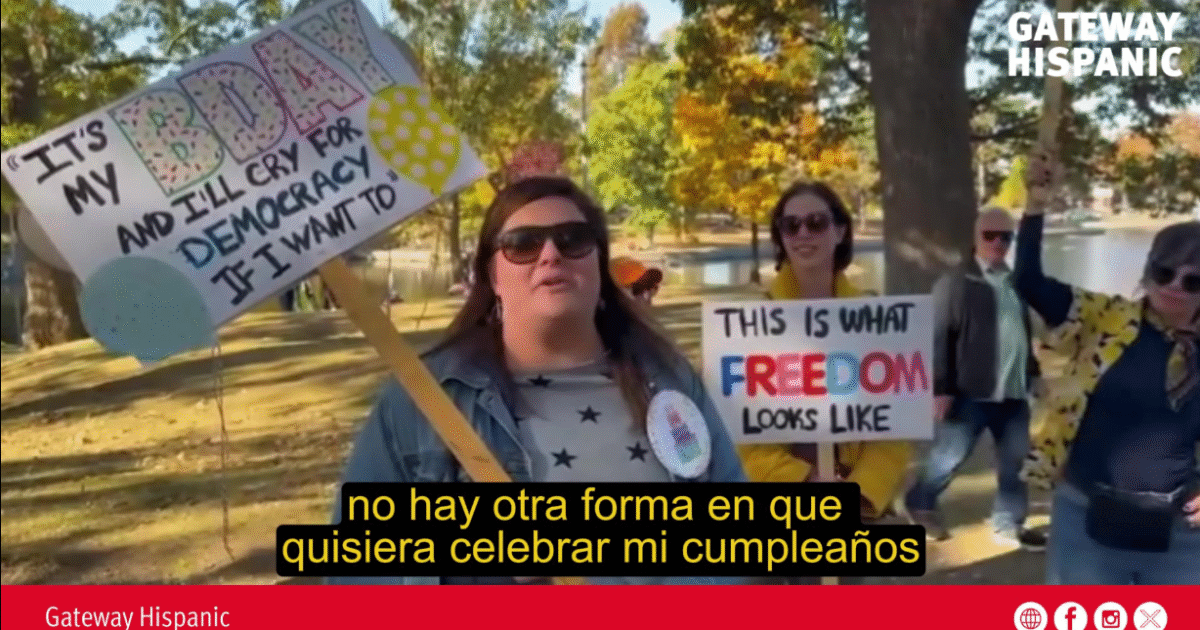 Participants at a birthday celebration rally hold signs advocating for democracy and freedom in a park setting, surrounded by autumn foliage.