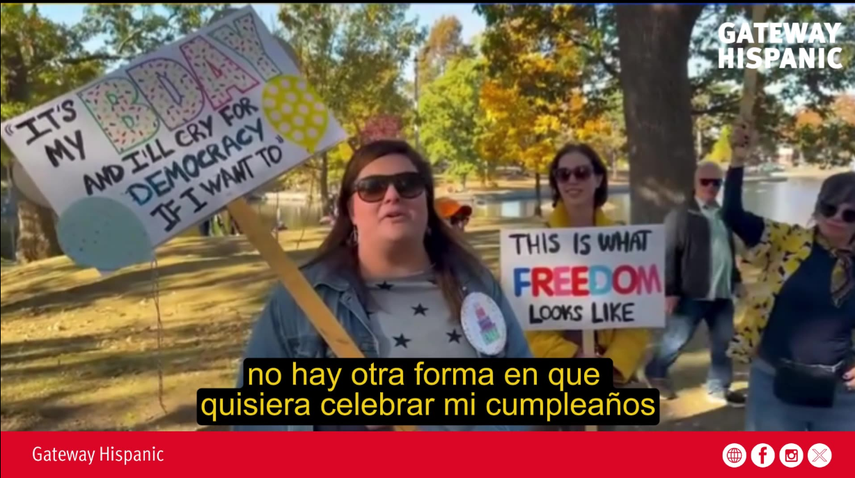 Participants at a birthday celebration rally hold signs advocating for democracy and freedom in a park setting, surrounded by autumn foliage.