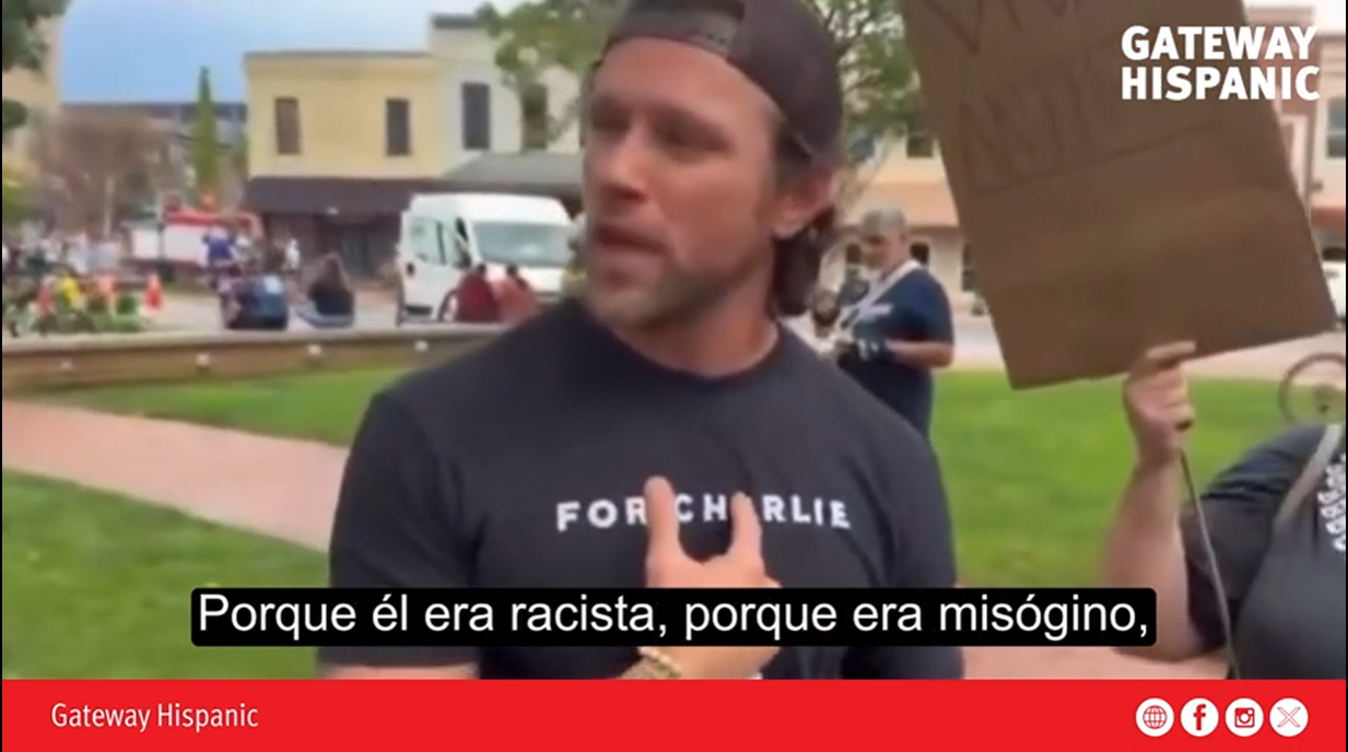 Man speaking passionately at a protest, addressing issues of racism and misogyny, with a crowd and signs in the background.