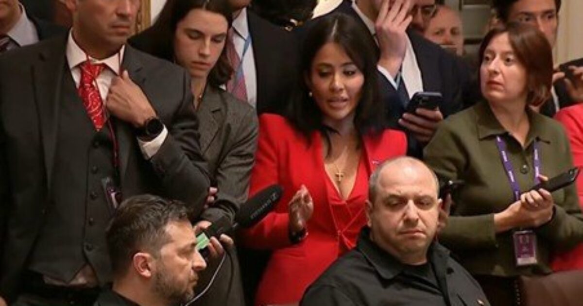 A diverse group of people in formal attire, including a woman in a red dress speaking, gathered at a press event or conference.