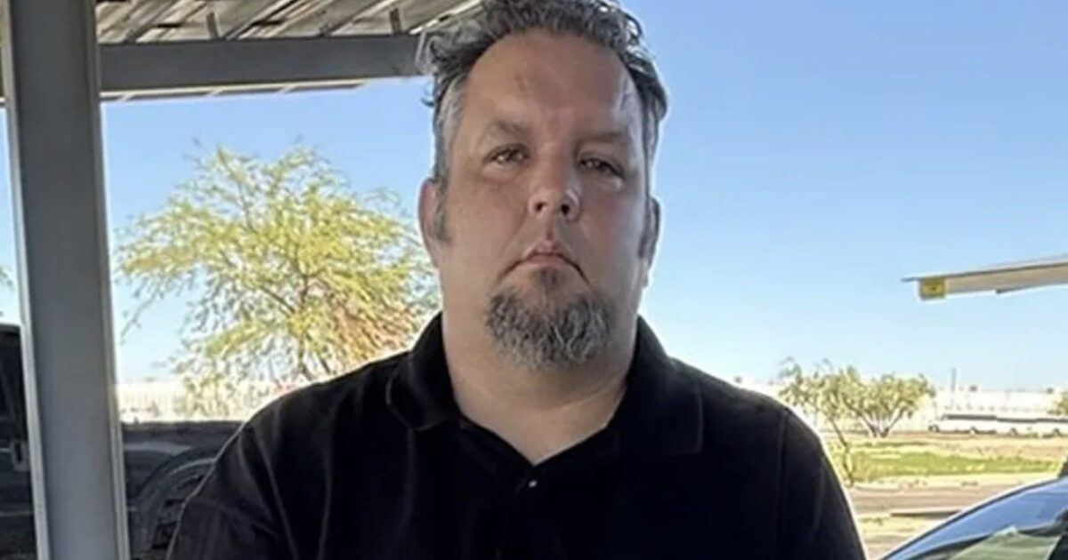 Man with a goatee standing outdoors under a shaded area, with a clear blue sky and trees in the background.