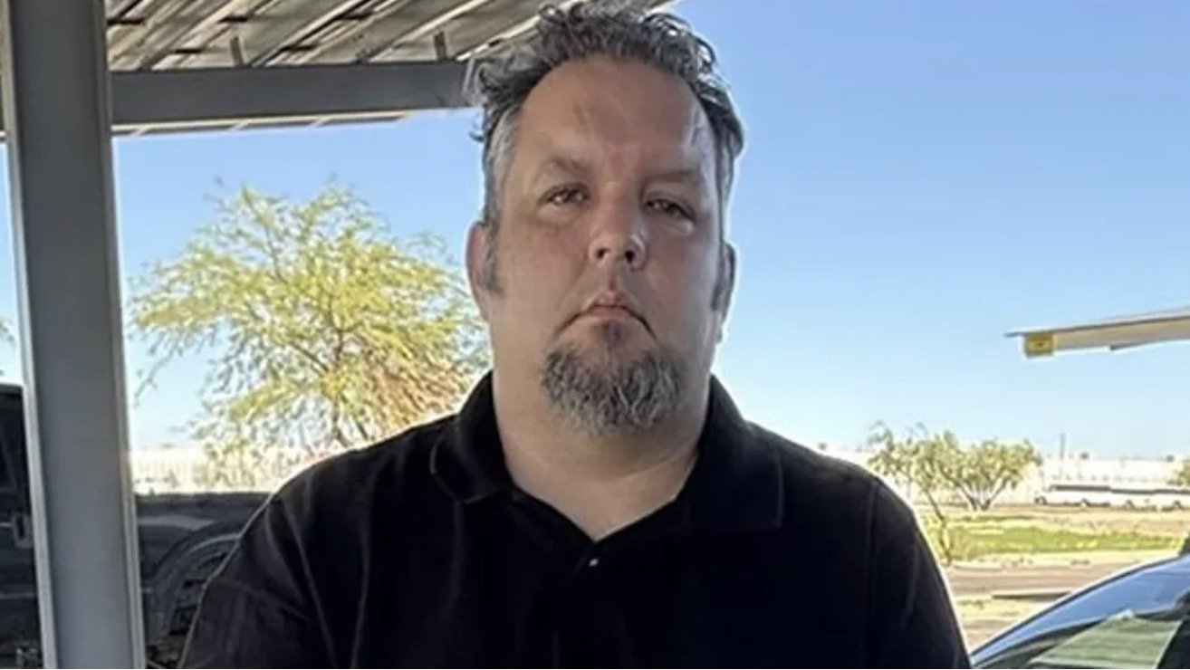 Man with a goatee standing outdoors under a shaded area, with a clear blue sky and trees in the background.